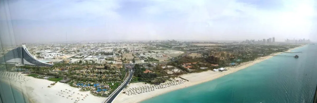 An elevated view along a long beach lined with parasols, greenery and a road beside it