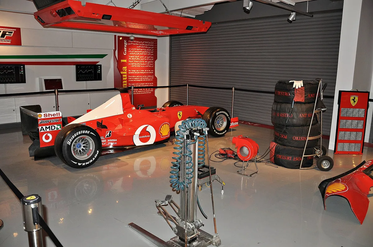 A red Formula One car inside a covered display garage, with stacked racing tyres and pit equipment beside it