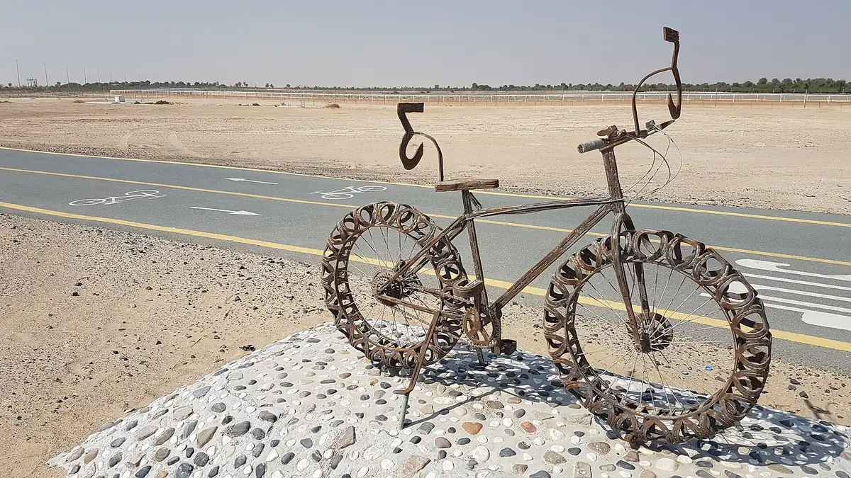 A rusted metal bicycle sculpture on a pebble mound beside a marked cycle lane crossing open desert