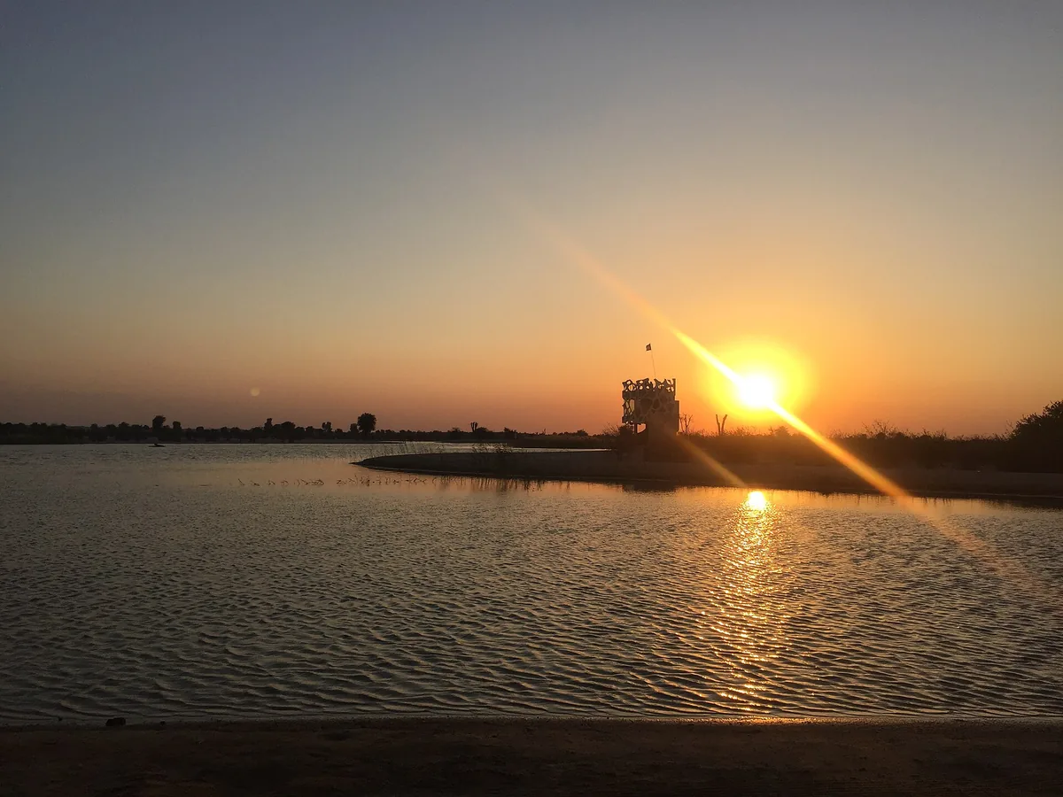 A wide desert lake with still water, the sun dropping behind the far bank and laying a trail of light across the surface