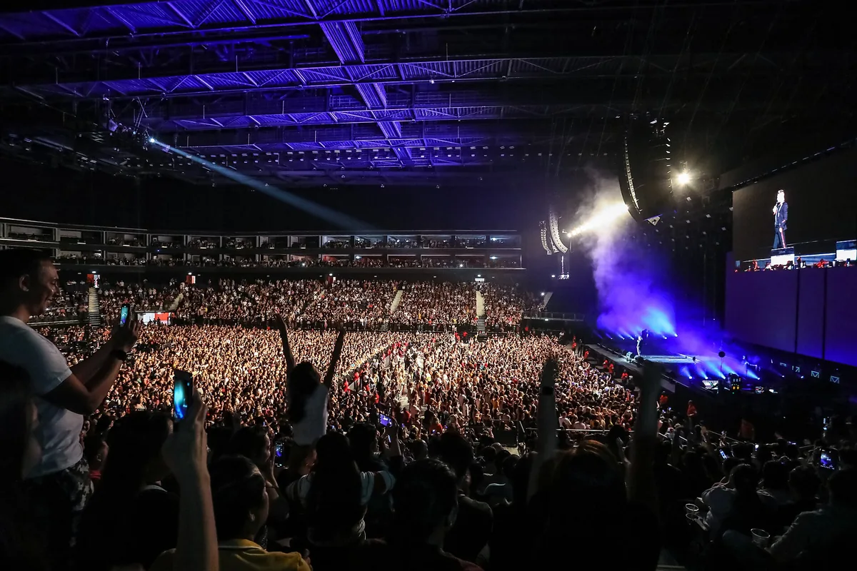 A packed indoor arena with blue beams of light above the stage