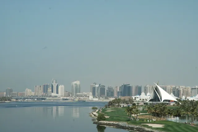 A green golf course beside a creek, a white sail shaped building and a skyline behind