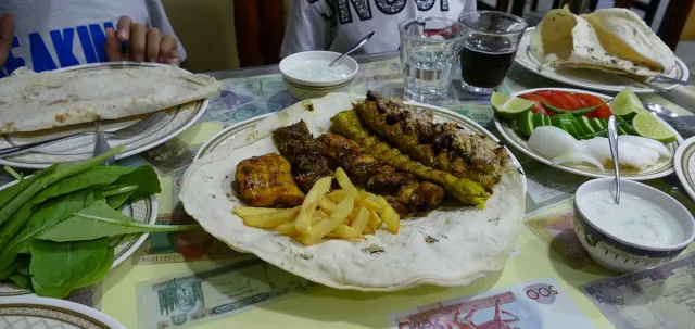 A plate of grilled meat skewers and chips on flatbread beside salad and drinks