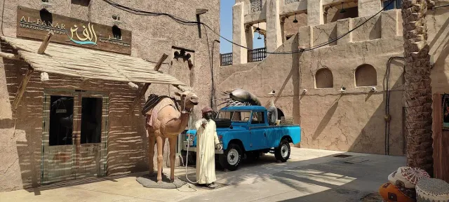 A traditional lane with a mud wall, a camel, an old blue pickup and a wind tower