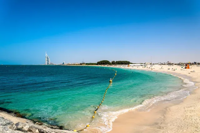 A curving sandy beach facing turquoise water with a sail shaped hotel on the horizon