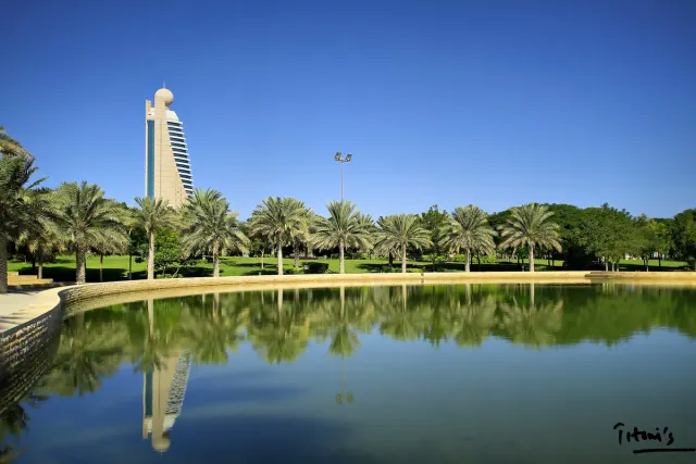 A lake reflecting palms and a tall tower with green lawns and a blue sky