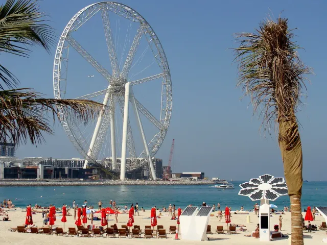 A giant white wheel across the water and a beach with red parasols and palms